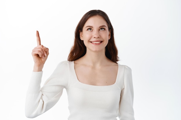 Beautiful caucasian girl model pointing fingers up looking at top advertisement with dreamy pleased smile showing good promo deal standing over white background