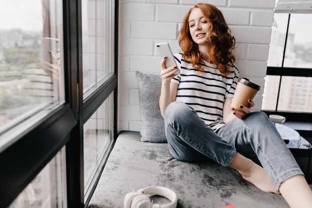Free photo beautiful caucasian girl with phone drinking coffee carefree redhaired woman enjoying latte