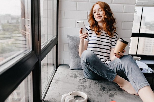 Bella ragazza caucasica con il telefono che beve caffè donna dai capelli rossi spensierata che gode del latte