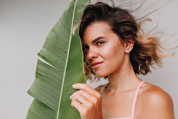 Beautiful caucasian girl with tanned skin touching her face with green leaf. Indoor photo of charming curly female model hiding behind exotic plant.