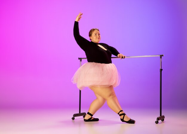 네온 불빛에 그라데이션 퍼플 핑크 스튜디오 배경에 발레 댄스를 연습하는 아름다운 백인 플러스 사이즈 모델. 동기 부여, 포용, 꿈 및 업적의 개념. 발레리나가 될만한 가치가 있습니다.