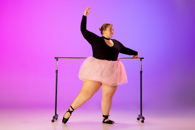 Beautiful caucasian plus size model practicing ballet dance on gradient purple-pink studio background in neon light. concept of motivation, inclusion, dreams and achievements. worth to be ballerina.