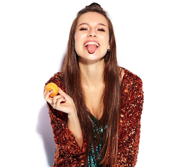 Free photo beautiful caucasian smiling hipster brunette woman model in bright shinny reflecting summer stylish jacket and green dress isolated . eating french macaroon and showing her tongue