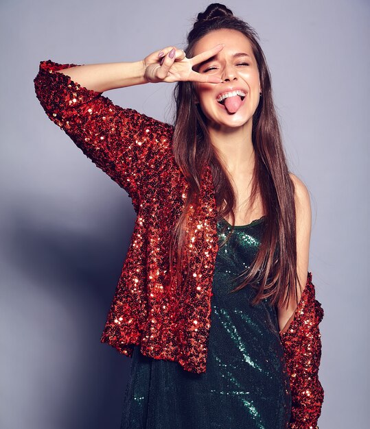 Beautiful caucasian smiling hipster brunette woman model in bright shinny reflecting summer stylish jacket and green dress posing on gray . Showing peace sign