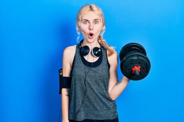 Beautiful caucasian sports woman with blond hair wearing sportswear using dumbbells scared and amazed with open mouth for surprise disbelief face