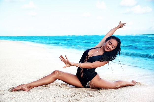 Beautiful caucasian sunbathed woman model with dark long hair in black elastic swimsuit for yoga doing split and posing on summer beach with white sand on blue sky and ocean