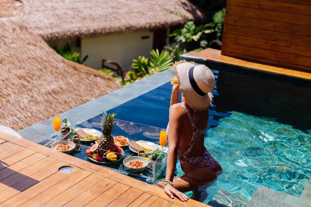 Beautiful caucasian tanned woman in bikini and straw hat with floating breakfast at amazing luxury bali style villa at sunny day by swimming pool, tropical background.