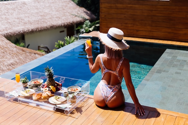 Beautiful caucasian tanned woman in bikini and straw hat with floating breakfast at amazing luxury bali style villa at sunny day by swimming pool, tropical background.