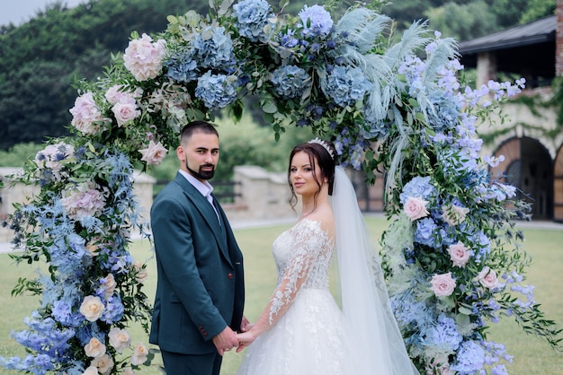 Free photo beautiful caucasian wedding couple is standing in front of decorated with blue hydrangea archway and holding hands together