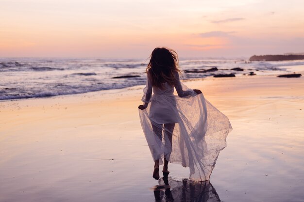 Beautiful caucasian woman alone in white dress at purple sunset by ocean