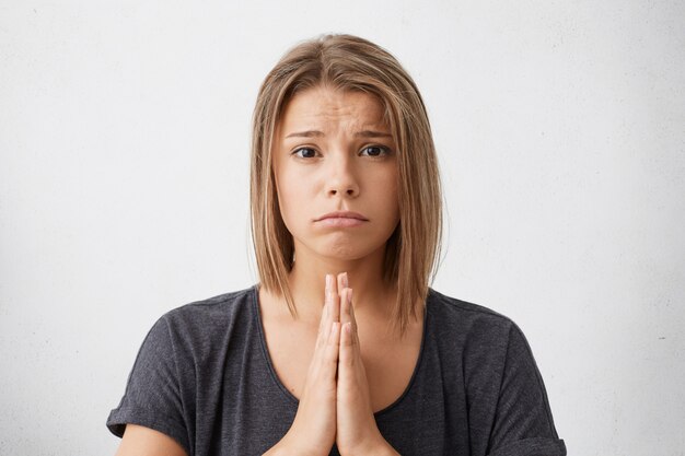 Beautiful Caucasian woman charming eyes and sorrowed look holding palms together begging for good mark. Sad pretty girl praying for good faith.