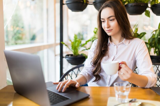 Beautiful Caucasian woman dreaming about something while sitting with portable net-book in modern cafe bar. Young charming female freelancer thinking about new ideas during work on laptop computer