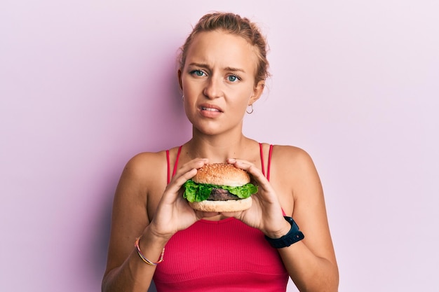 Beautiful caucasian woman eating a tasty classic burger clueless and confused expression doubt concept