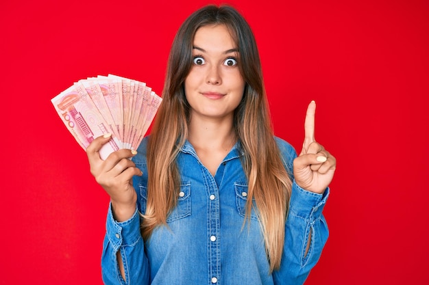 Beautiful caucasian woman holding yuan chinese banknotes smiling with an idea or question pointing finger with happy face, number one