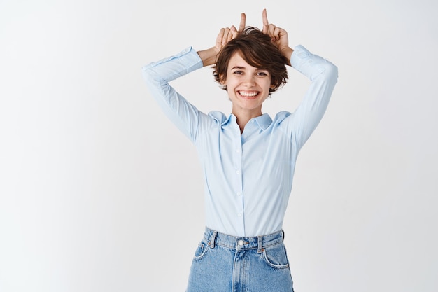 Free photo beautiful caucasian woman in office clothings, showing horns on head and smiling, standing on white wall