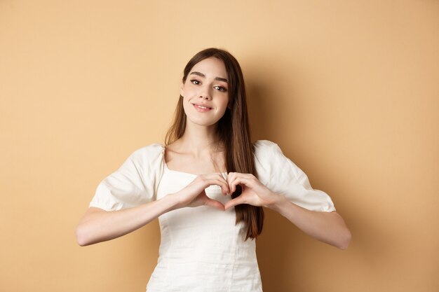 Beautiful caucasian woman say i love you showing heart gesture and smile at camera beige background ...