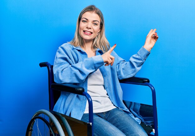 Красивая кавказская женщина, сидящая на инвалидной коляске, улыбается и смотрит в камеру, указывая двумя руками и пальцами в сторону