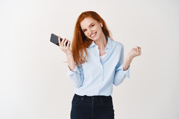 Beautiful caucasian woman with red hair listening music in wireless headphones, holding smartphone and smiling at front, white wall