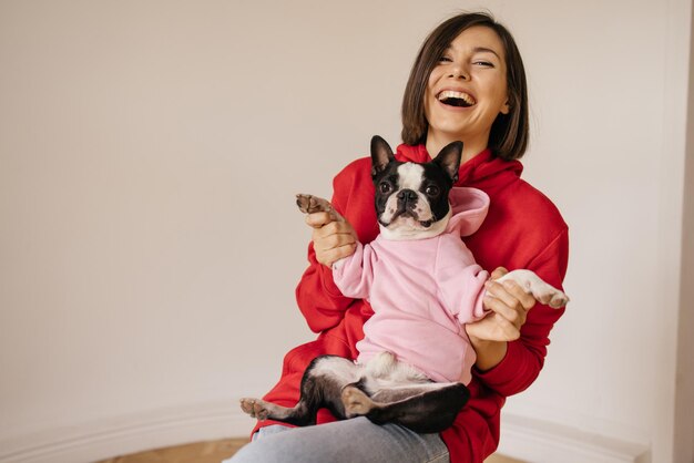 Beautiful caucasian young girl laughs while holding boston terrier dog on her knees in white background Lifestyle and friendship concept