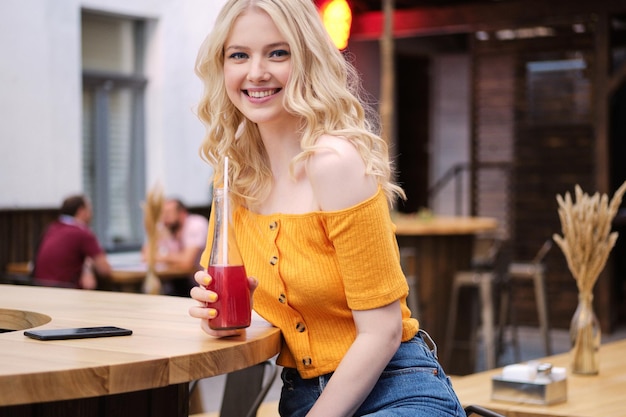Beautiful charming blond girl happily looking in camera with lemonade in courtyard of city cafe