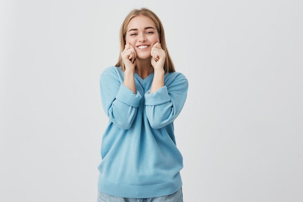 Beautiful, charming european female with straight fair hair wearing blue sweater and jeans looking  with smile and pinching her cheeks. Pretty, playful girl posing.