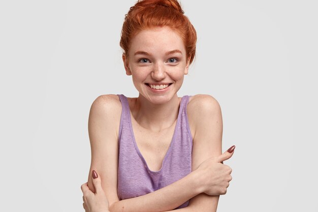 Beautiful charming young red haired lady embraces herself, feels pleased and shy to be invited on party, wears casual purple t shirt, looks directly, models against white wall