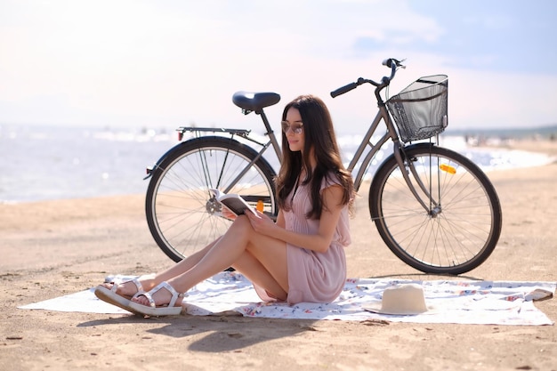 Красивая веселая брюнетка в шляпе наслаждается летним временем, читая книгу на море рядом со своим велосипедом.