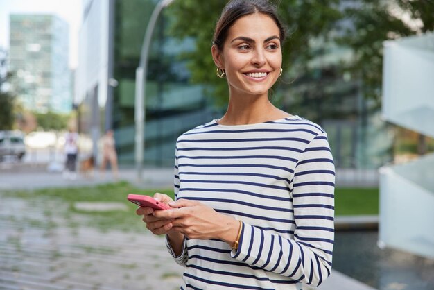 Beautiful cheerful brunette woman wears striped jumper concentrated away uses mobile phone for chatting online poses against blurred city background. Positive female tourist tries to find route