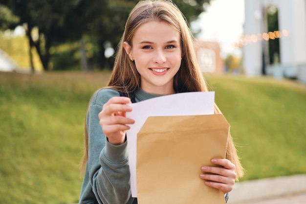 都会の公園でカメラを見て幸せに試験結果の封筒を開く美しい陽気なカジュアルな学生の女の子
