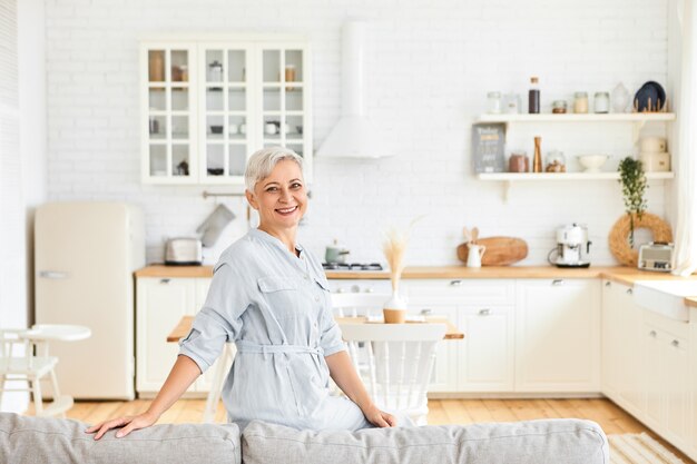 Beautiful cheerful Caucasian housewife in elegant dress posing in living room, sitting on back of large gray couch, smiling, going to show you her spacious cozy apartment. Aged people and lifestyle