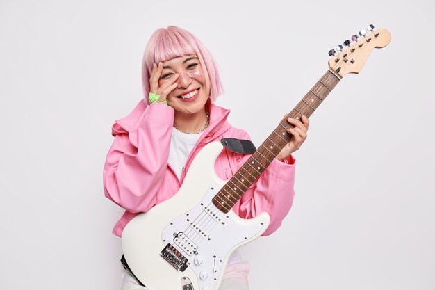 Beautiful cheerful female musician plays electric guitar being member of popular rock group has pink hairstyle wears jacket 