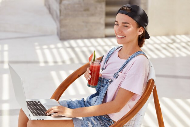 Beautiful cheerful female in trendy cap, dressed in fashionable clothing, drinks fresh smoothie, searches information on laptop computer