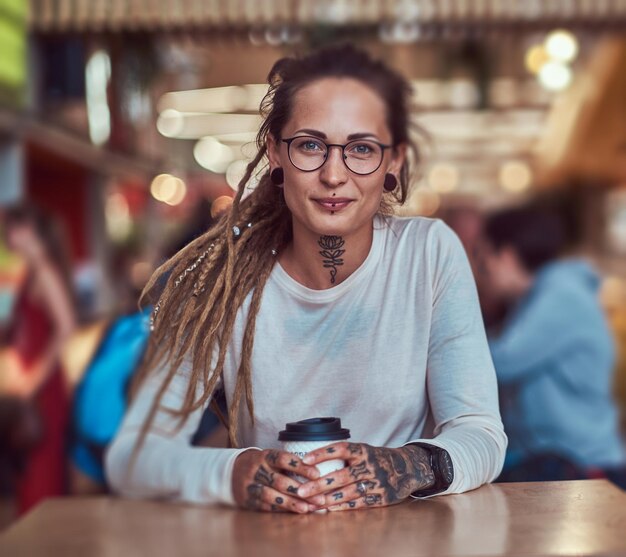 入れ墨とドレッドヘアを持つ美しい陽気な女の子は、コーヒーを飲みながらフードコートに座っています。