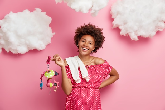 Beautiful cheerful pregnant woman prepares for motherhood, has big belly, buys toys and clothes for unborn child, enjoys happy expectations, isolated on pink wall with white clouds above