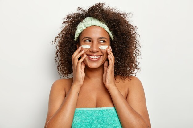 Free photo beautiful cheerful woman applies cosmetics face care cream, looks gladfully aside, wears headband, stands wrapped in towel, cares about appearance and beauty