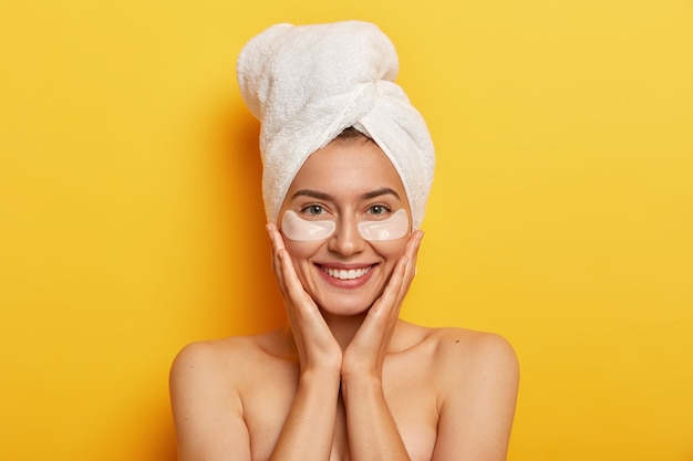 Beautiful cheerful woman has wrappped towel for drying hair, keeps both hands on cheeks, smiles gently, has fresh skin, cares about complexion, wears patches under eyes