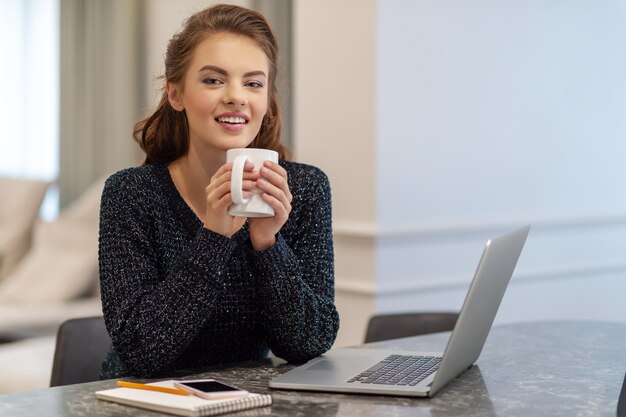 コーヒーカップを持って、自宅のキッチンで笑顔で目をそらす美しい陽気な若い女性