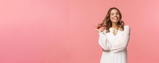 Beautiful and confident blond young 20s woman in stylish white cute dress touching hair and smiling satisfied getting dressed for romantic date concept of women and beauty pink background