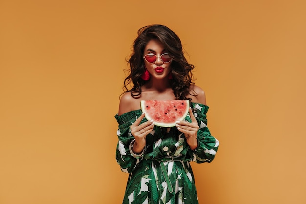 Beautiful cool girl with curly dark hair in trendy bright summer dress and red sunglasses posing with watermelon on orange backdrop