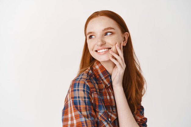 Beautiful coquettish redhead girl smiling with white perfect teeth, touching pale face without makeup and looking left happy, standing over white wall
