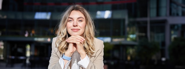 Free photo beautiful corporate woman looks dreamy and smiles stands outside on street leans her head on hands