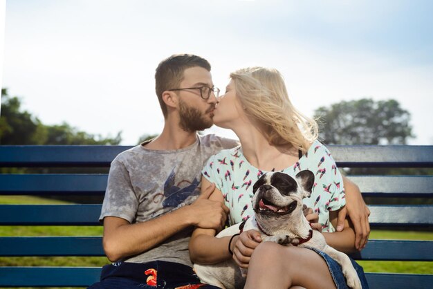 Beautiful couple kissing, walking, sitting with French bulldog on bench in park. Outdoor.