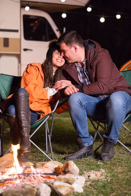 Beautiful couple recreating along campfire in the mountains with their retro camper van in the background.