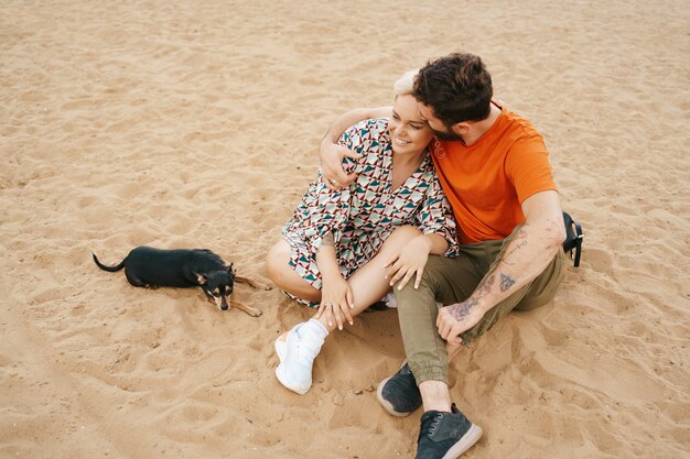 Beautiful couple relaxing on sand hugging and kissing while playing with their positive dog