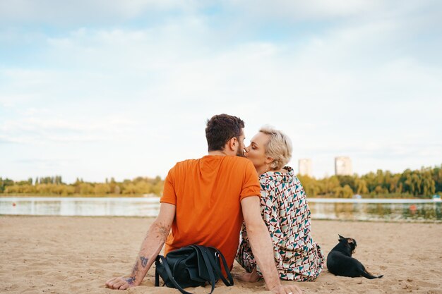 Красивая пара расслабляется на песке, обнимается и целуется, играя со своей позитивной собакой