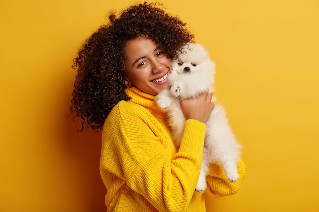 Beautiful curly Afro American lady in yellow oversized sweater, plays with favorite pet indoor, has happy mood, feels proud of having nice animal.