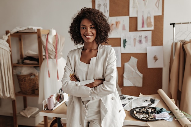 Beautiful curly brunette dark-skinned fashion designer poses in office, leans on table