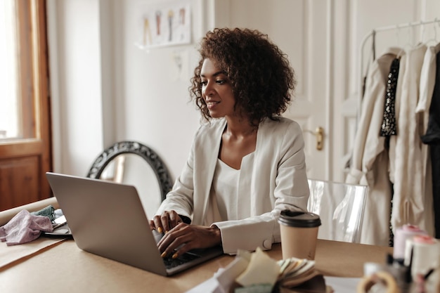 Бесплатное фото Красивая кудрявая темнокожая женщина в белой куртке смотрит на экран ноутбука, сидит за столом и улыбается в уютном офисе