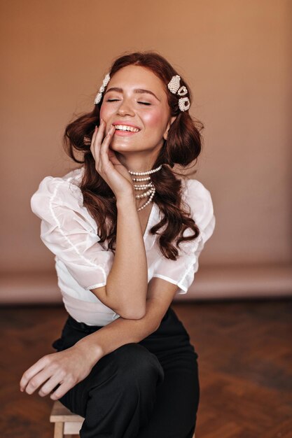 Beautiful curly girl dressed in white blouse and black pants sits on chair Woman with hairpins and pearl necklace posing in beige room