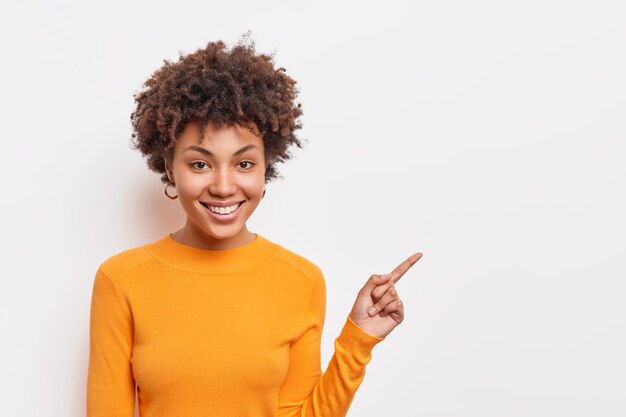美しい巻き毛のアフロアメリカ人女性は、空白のコピースペースのリンクポイントをクリックすることを提案し、白い壁の上に立つ方法が基本的なオレンジ色のジャンパーを着ていることを示しています。人と広告の概念